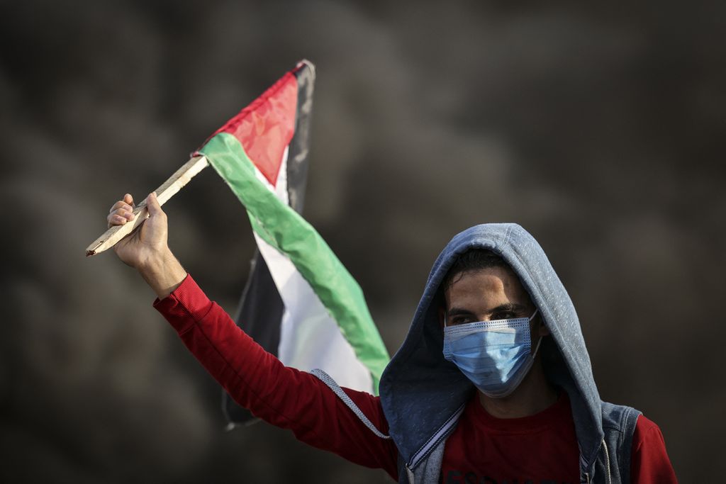 Seorang pengunjung-rasa mengibarkan bendera Palestina di tengah kepulan asap dari roda yang dibakar pada demontras idi sepanjang perbatasan dengan Israel di Kota Gaza, 25 Oktober 2022. (Photo by Mahmud HAMS / AFP)