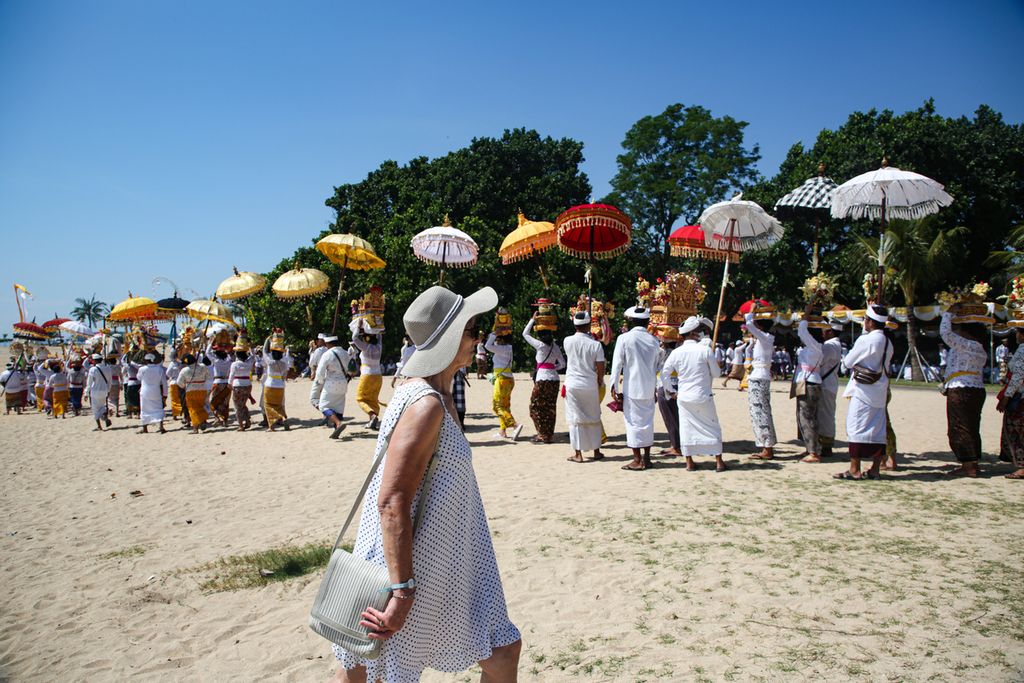 Turis asing melintas di dekat arak-arakan umat Hindu yang akan melaksanakan upacara Melasti di pantai Samuh, Badung, Bali, Minggu (19/03/2023).