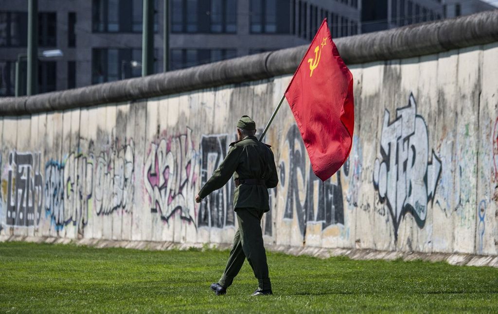 Seorang pria yang membawa bendera Soviet berjalan melewati bagian Tembok Berlin saat peringatan 60 tahun pembangunan Tembok Berlin, Jumat (13/8/2021).