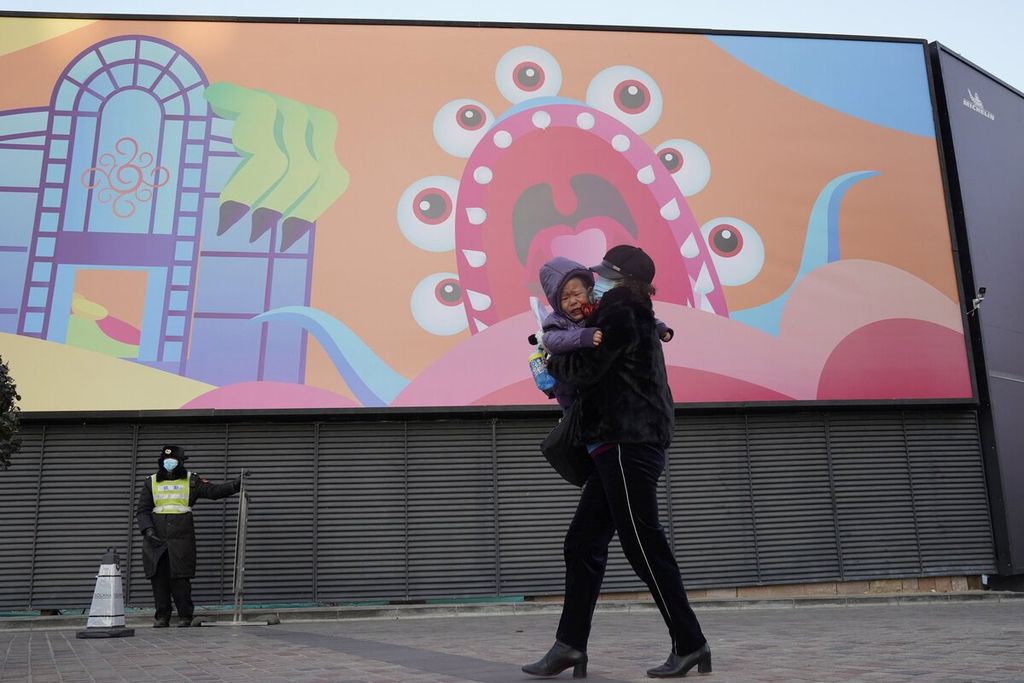 Seorang wanita menggendong anaknya yang menangis melewati sebuah mal yang kosong saat cuaca dingin dan angin kencang melanda Beijing, Rabu (6/1/2021). China dilaporkan tengah berjibaku menahan penyebaran wabah Covid-19 lewat galur Omicron. 
