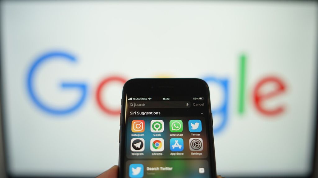 Laman pencarian Spotlight di ponsel yang menjalankan sistem operasi Apple iOS 14, di depan sebuah layar komputer yang menunjukkan logo Google.