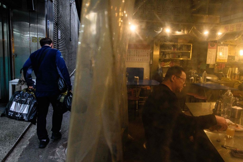 Seorang warga tengah menikmati makan malam di warung pinggir jalan, saat seorang pekerja tengah membawa barang dalam bungkusan plastik melintas di belakangnya di Pasar Namdaemun, Seoul pada Senin (13/3/2023).