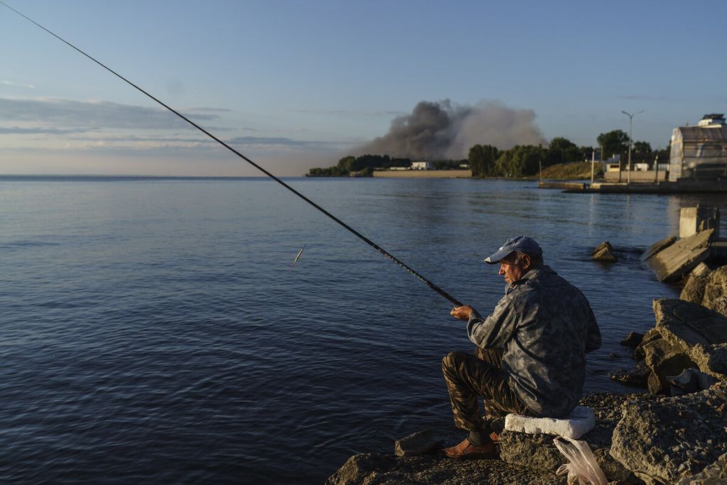 Warga memancing ikan di tepi pantai dengan latar asap mengepul setelah terjadi serangan dari Rusia pada instalasi militer Ukraina di Vyshhorod, Kyiv, Kamis (28/7/2022). Sebagian kecil warga yang tidak mengungsi tetap melanjutkan aktivitas sehari-hari di tengah peperangan.